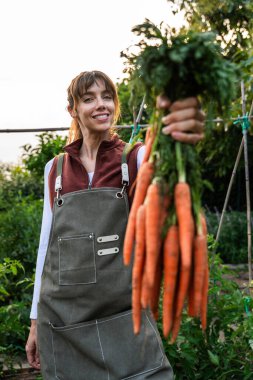 Gülümseyen dişi çiftçi canlı bir meyve bahçesinde taze toplanmış havuçları gururla sergiliyor, çiftlikten sofraya tarımın ve sağlıklı beslenmenin özünü somutlaştırıyor.