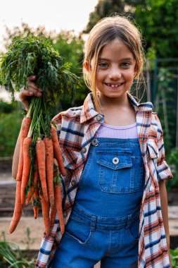 Gülümseyen genç çiftçi kız canlı bir bahçede taze toplanmış havuçları gururla tutuyor sürdürülebilir tarımı ve sağlıklı beslenmeyi kutluyor.