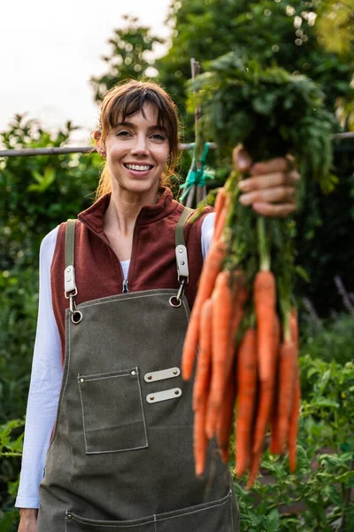 Gülümseyen kadın çiftçi elinde taze toplanmış havuçlarla, sağlıklı beslenmeyi kutluyor ve canlı ürünlerle çevrili biyolojik çiftliğinde sürdürülebilir tarımı destekliyor.