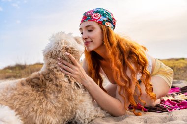 Renkli bir bandana takan kızıl saçlı kadın bej karalamalarını güneşli kumda sarılıyor. Şefkatli, rahat bir arkadaşlık anını ve sahilde yaz neşesini paylaşıyor.
