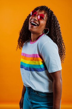 Gökkuşağı tişörtü ve kırmızı güneş gözlüğü giyen neşeli genç bir kadının iki eli turuncu arka planda barış işareti gösteren bir stüdyo fotoğrafı.