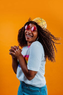 Genç kaygısız Latin kadın gökkuşağı tişörtü, komik şapka ve pembe gözlük takıyor turuncu arka planda poz veriyor, kıvırcık saçları var ve gülümsüyor.
