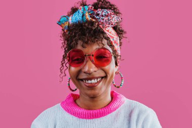 Başörtüsü, renkli küpeler, pembe güneş gözlükleri ve pembe bir arka planda gülümseyen fuşya ve beyaz süveter giyen kıvırcık saçlı neşeli genç bir kadının stüdyo portresi.