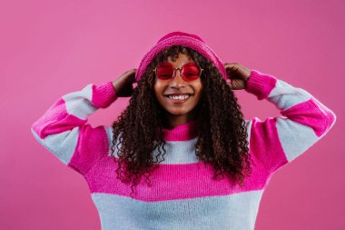 Pembe ve beyaz çizgili bir süveter, pembe güneş gözlüğü ve pembe bir şapka takan neşeli bir mankenin atölye çekimi.