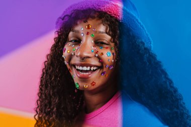 Stüdyo portresi kıvırcık saçlı, yüzünde renkli elmaslar olan, pembe bir şapka ve turuncu bir tişört giyen, canlı, renkli bir arka plana poz veren neşeli bir genç kadın.