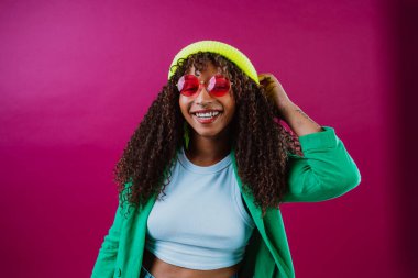 Kıvırcık saçlı, parlak yeşil ceketli, açık mavi bluzlu, sarı yün şapkalı ve pembe gözlüklü neşeli genç bir kadın canlı bir fuşya arka planına gülümsüyor.