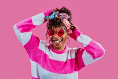 Başörtüsü, renkli küpeler, pembe güneş gözlükleri ve pembe bir arka planda gülümseyen fuşya ve beyaz süveter giyen kıvırcık saçlı neşeli genç bir kadının stüdyo portresi.