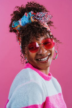 Başörtüsü, renkli küpeler, pembe güneş gözlükleri ve pembe bir arka planda gülümseyen fuşya ve beyaz süveter giyen kıvırcık saçlı neşeli genç bir kadının stüdyo portresi.