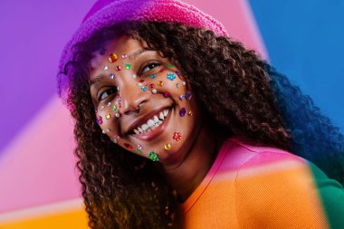 Stüdyo portresi kıvırcık saçlı, yüzünde renkli elmaslar olan, pembe bir şapka ve turuncu bir tişört giyen, canlı, renkli bir arka plana poz veren neşeli bir genç kadın.
