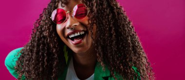 Yeşil ceket ve pembe güneş gözlüğü takan neşeli genç bir müdürün stüdyo portresi, fuşya arka planında gülüyor, mutluluğu ve başarıyı ifade ediyor.