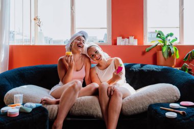 Kafalarında havluları ve göz altı bantları olan iki neşeli kadın rahat bir koltukta oturuyor, gülüyor ve güzellik ürünleri tutuyor, rahatlatıcı bir spa gününün tadını çıkarıyorlar.