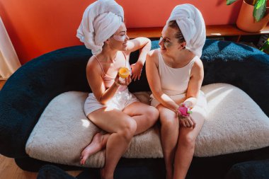 İki kadın evlerinde rahat bir spa gününün tadını çıkarıyorlar. Kafalarına havlular sarılmış ve göz altı bantları uygulanmış, çeşitli güzellik ürünleriyle çevrili rahat bir koltukta oturuyorlar.