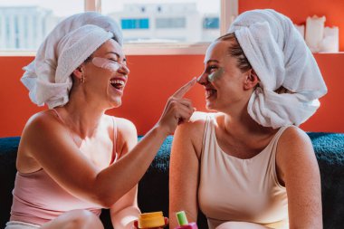 Kafalarına havlu sarılmış iki neşeli kadın bir kanepede oturmuş kozmetik ürünler uyguluyor ve rahatlatıcı bir spa gününün tadını çıkarıyorlar.