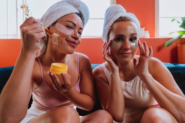 Havluya sarılmış, kozmetik ürünler ve göz bantları yapıştıran iki arkadaş, güzellik rutinlerini yaparken nefis bir kişisel bakım ve rahatlama anının tadını çıkarıyorlar.