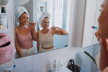 Kafalarına havlu geçirmiş iki neşeli kadın, modern bir banyoda sabah güzelliklerinin keyfini çıkarırken dişlerini fırçalarlar ve altın göz bantları takarlar.