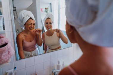 Kafalarına havlu geçirmiş iki neşeli kadın, modern bir banyoda sabah güzelliklerinin keyfini çıkarırken dişlerini fırçalarlar ve altın göz bantları takarlar.