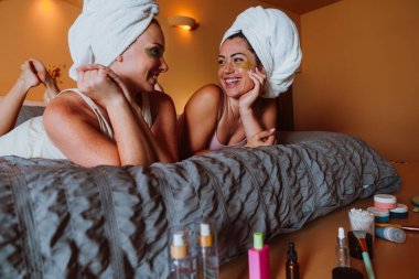 Kafalarında havluları olan iki gülümseyen kadın yataklarında uzanmış gözlerinin altına kozmetik bantlar takmış rahat bir yatak odasında rahatlatıcı bir cilt bakımı ve arkadaşlığın tadını çıkarıyorlar.