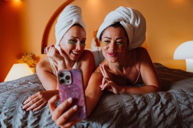 Havluya sarılmış ve göz altı bantları takmış iki neşeli kadın, rahatlatıcı bir cilt bakımı gecesinde bir yatağa uzanır, gülümser ve birlikte akıllı telefondan keyif alırlar.