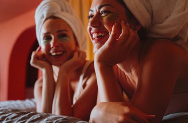 İki neşeli kadın, göz altı bantlı havlulara sarılmış, bir yatakta uzanmış, evde rahatlatıcı bir spa gününün tadını çıkarırken, kendine bakıcılık ve cilt bakımı yapıyor.