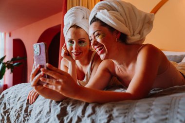 Havluya sarılmış ve göz altı bantları takmış iki neşeli kadın, rahatlatıcı bir cilt bakımı gecesinde bir yatağa uzanır, gülümser ve birlikte akıllı telefondan keyif alırlar.