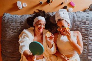 İki neşeli kadın kafalarına ve göz altı bantlarına sarılmış havlularla yatakta uzanıyor, bir ayna tutuyor ve selfie çekiyor, evde rahatlatıcı bir spa gününün tadını çıkarıyorlar.