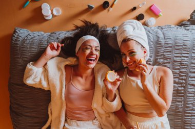 Göz altı bantları ve bornozları olan iki neşeli genç kadın yatağa uzanıyor, güzellik ürünleri uyguluyor, eğleniyor ve cilt bakımları sırasında barış işareti yapıyorlar.