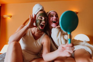 İki neşeli bayan arkadaş evlerinde rahatlatıcı bir spa günü geçiriyorlar, kafalarına bornoz ve havlu geçiriyorlar, yüz kozmetik maskeleri takıyorlar ve kendilerine küçük bir aynada bakıyorlar.