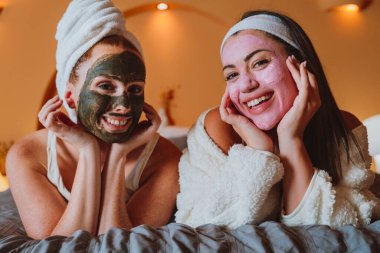 İki neşeli kadın evde rahatlatıcı bir spa gününün tadını çıkarıyorlar, kozmetik maske ve bornozlarla dolu bir yatakta uzanıyorlar, özgüven ve refahı kucaklıyorlar.