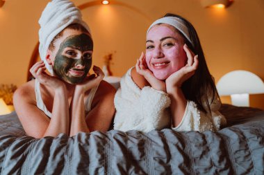 İki neşeli kadın evde rahatlatıcı bir spa gününün tadını çıkarıyorlar, kozmetik maske ve bornozlarla dolu bir yatakta uzanıyorlar, özgüven ve refahı kucaklıyorlar.