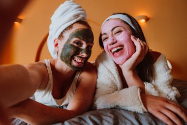 İki neşeli kadın evde rahatlatıcı bir spa gününün tadını çıkarıyorlar, kozmetik maske ve bornozlarla dolu bir yatakta uzanıyorlar, özgüven ve refahı kucaklıyorlar.