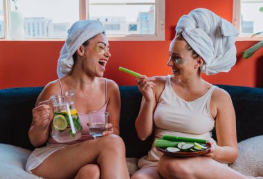 Kafasında havlu ve göz bandı olan iki kadın bir kanepeye oturmuş, bir sürahide detoks suyu döküyor, kereviz ve salatalık yiyor, sağlıklı bir yaşam tarzını teşvik ediyorlar.