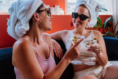 Kafalarına havlu geçirmiş gülümseyen iki arkadaş bir kanepede oturmuş zeytinli kokteyllerin tadını çıkarıyor ve kadeh kaldırıyor ve birlikte rahatlatıyorlar.