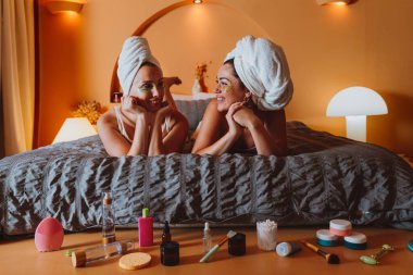 Kafalarında havluları olan iki gülümseyen kadın yataklarında uzanmış gözlerinin altına kozmetik bantlar takmış rahat bir yatak odasında rahatlatıcı bir cilt bakımı ve arkadaşlığın tadını çıkarıyorlar.