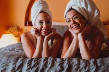 İki neşeli kadın, göz altı bantlı havlulara sarılmış, bir yatakta uzanmış, evde rahatlatıcı bir spa gününün tadını çıkarırken, kendine bakıcılık ve cilt bakımı yapıyor.
