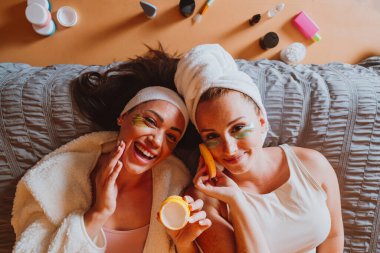 Göz altı bantları ve bornozları olan iki neşeli genç kadın yatağa uzanıyor, güzellik ürünleri uyguluyor, eğleniyor ve cilt bakımları sırasında barış işareti yapıyorlar.