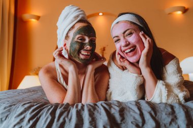 İki neşeli kadın evde rahatlatıcı bir spa gününün tadını çıkarıyorlar, kozmetik maske ve bornozlarla dolu bir yatakta uzanıyorlar, özgüven ve refahı kucaklıyorlar.