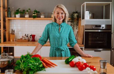 Modern mutfağında ayakta duran mutlu olgun bir kadın tezgahta çeşitli taze sebzelerle sağlıklı bir yemek hazırlamaya, sağlıklı bir yaşam tarzını desteklemeye hazır.