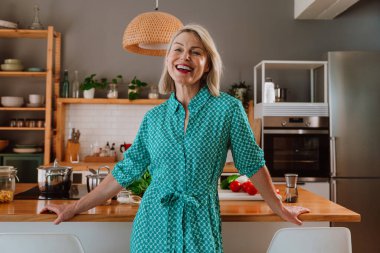 Modern mutfağında ayakta duran mutlu olgun bir kadın tezgahta çeşitli taze sebzelerle sağlıklı bir yemek hazırlamaya, sağlıklı bir yaşam tarzını desteklemeye hazır.