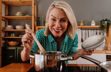 Mutlu olgun bir kadın ahşap bir kaşıkla yemek pişirirken ve modern bir mutfakta tencere kapağı tutarken gülümsüyor. Lezzetli bir ev yemeği hazırlama sürecinin tadını çıkarıyor.