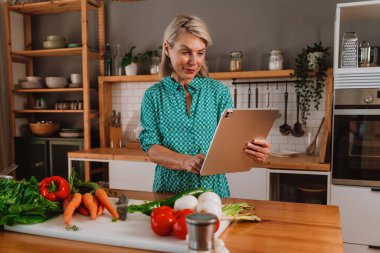 Dijital tablet kullanan gülümseyen olgun bir kadın ev mutfağında sağlıklı yemek için taze malzemeler hazırlarken, yemek pişirmekten ve modern teknolojiden zevk alır.
