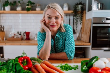 Taze sebzelerle dolu bir mutfak masasına yaslanıp sağlıklı beslenmeyi ve dengeli bir yaşam tarzını kucaklamayı kutlayan mutlu olgun bir kadın.