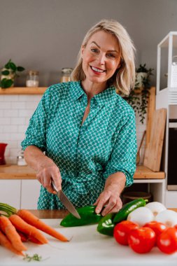 Yeşil elbise giyen gülümseyen yaşlı kadın modern bir mutfakta taze sebzeleri kesiyor sağlıklı beslenmeyi ve aktif yaşlanmayı somutlaştırıyor.