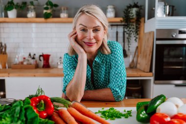 Taze sebzelerle dolu bir mutfak masasına yaslanıp sağlıklı beslenmeyi ve dengeli bir yaşam tarzını kucaklamayı kutlayan mutlu olgun bir kadın.