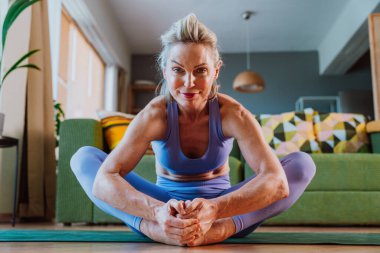 Oturma odasında yoga pratiği yapan, yoga minderinde bağlı bir pozisyonda oturan, yaşlılıkta sağlıklı yaşam tarzını ve sağlığını destekleyen formda bir kadın.
