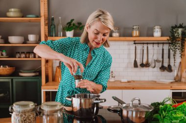 Modern mutfakta fırında yemek hazırlarken biberi paslanmaz çelik tencereye öğüten mutlu olgun kadın evde yemek pişirme hobisinin keyfini çıkarıyor.