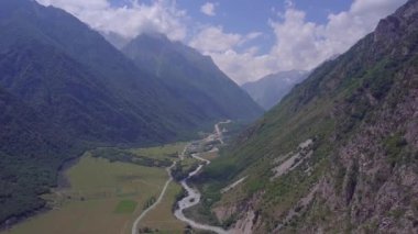 Kuzey Kafkasya 'nın devasa görkemli dağları. Vadinin dibinde çalkantılı bir nehir var. Hava görüntüsü. Güneşli bir gün. Dağlar, tepeler, uçurumlar, kayalar, tepeler, manzaralar, yeşil çimenler, gökyüzü, bulutlar