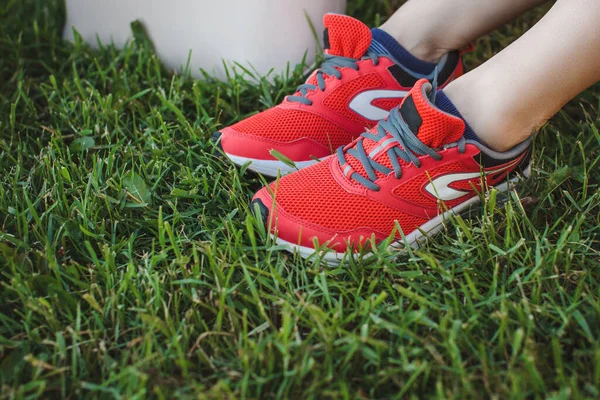 Girl in red sports sneakers sits resting on the grass in the park after jogging. Legs close up.