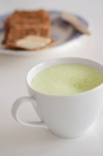 Υγιή πράσινο τσάι latte matcha με σόγια και βρώμη γάλα σε ένα φλιτζάνι τσαγιού λευκό διακοσμημένο με βασιλικό και αφρό στην κορυφή - ένα ζεστό ρόφημα να ενισχύσει τη λειτουργία του εγκεφάλου και ως πρόληψη καρκίνου! Φρυγανιά με το βούτυρο θολή σε φόντο — Φωτογραφία Αρχείου