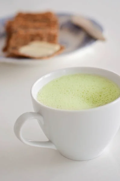 Egészséges zöld tea matcha latte a szója és a tej, bazsalikommal és a hab a tetején - egy meleg ital agyműködés fokozására, és, mint a rák megelőzésére díszített fehér tea csészében! Kenyér és a vaj elmosódott háttér — Stock Fotó