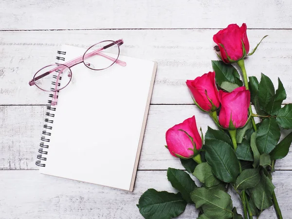 Blank note pad pink with eyeglases and red roses flower on white vintage wood background. Top view and flat lay style.